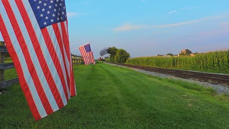 Ein-Niedriger-Blickwinkel-Auf-Eine-Reihe-Sanft-Wehender-Amerikanischer-Flaggen-Auf-Einem-Zaun-Bei-Ackerland,-Während-Sich-Während-Der-Goldenen-Stunde-Ein-Dampf-Personenzug-Nähert,-Der-Rauch-Bläst