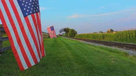 a low angle view of a line of gently waving american flags on a fence by farmlands as a steam passenger train blowing smoke approaches during the golden hour