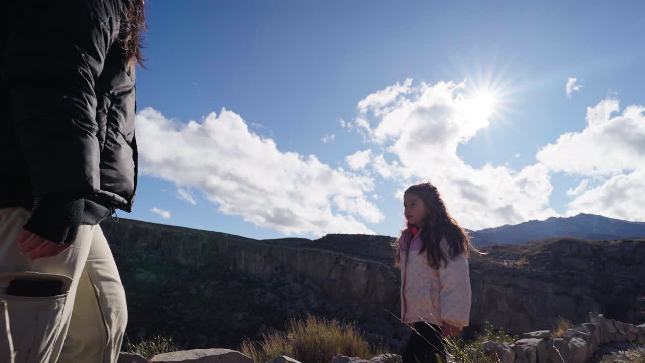 Static shot of an adult and child hugging on rocky terrain, facing a sunlit mountain landscape under a clear sky