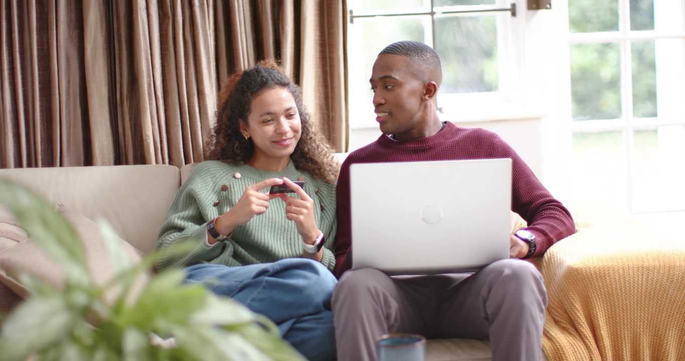 Couple on sofa in living room, woman tapping smartphone showing content while man adjusting laptop