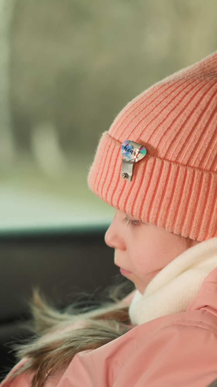 핑크색 베니와 재을 따뜻하게 입은 어린 소녀의 클로즈업, 차에 조용히 앉아, 그녀의 평온하고 평온한 표정, 그녀의 겨울 의상의 부드러움과 평화로운 순간을 강조합니다.