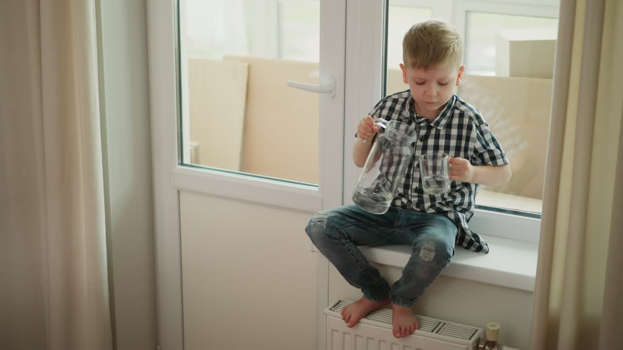 Fair skinned child wearing plaid shirt seated near window carefully pouring water from glass jar in right hand into glass cup in left hand lifting cup up and drinking