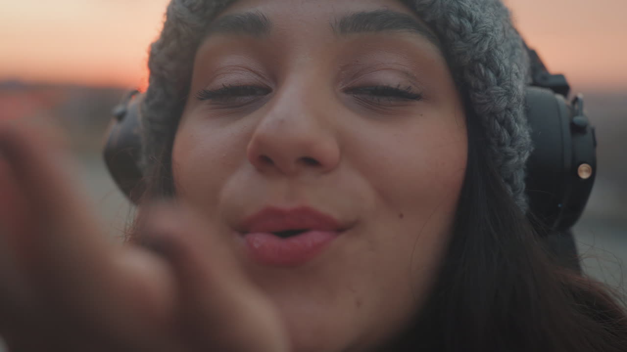 Closeup woman headphones blowing kiss toward camera, playful dancer vibe at rooftop sunset, warm golden glow on face, knitted beanie and puffy jacket, soft film grain, candid joyful expression, subtle