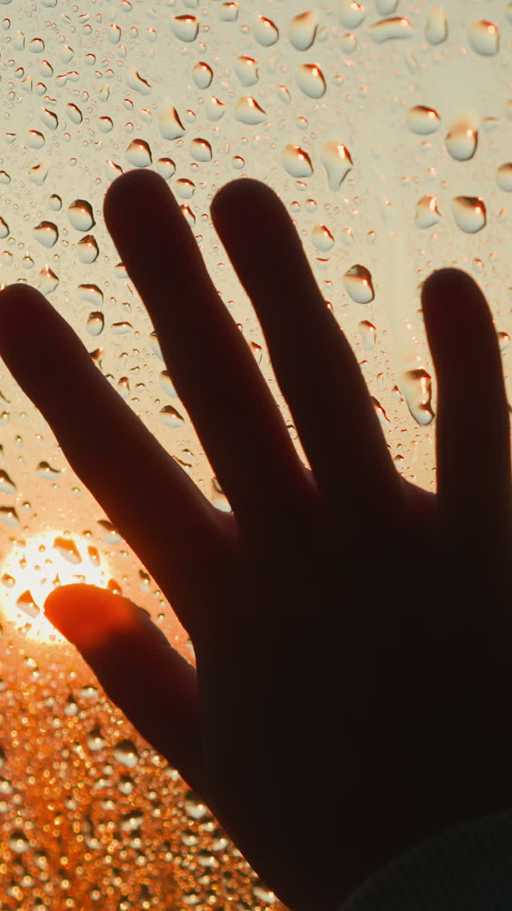 은 창문의 클로즈업에서 슬의 상징으로 손바닥 실루, 비가 오는 날 해가 지는 빛에 은 물방울이 있는 유리 위에 검은 손, 우울한 날씨