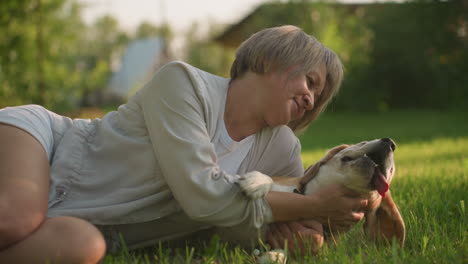 woman lying with her dog on grassy field, gently touching and holding her dog affectionately, both enjoying peaceful moment under warm sunlight, surrounded by lush greenery