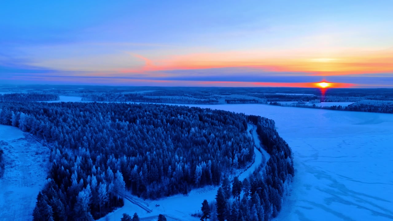 Vintersol som berører horisonten over frossen elv og skog, Lappland. Luftfoto av svingete vei gjennom snødekt taiga med oransje solstråler ved iskanten av en innsjø.
