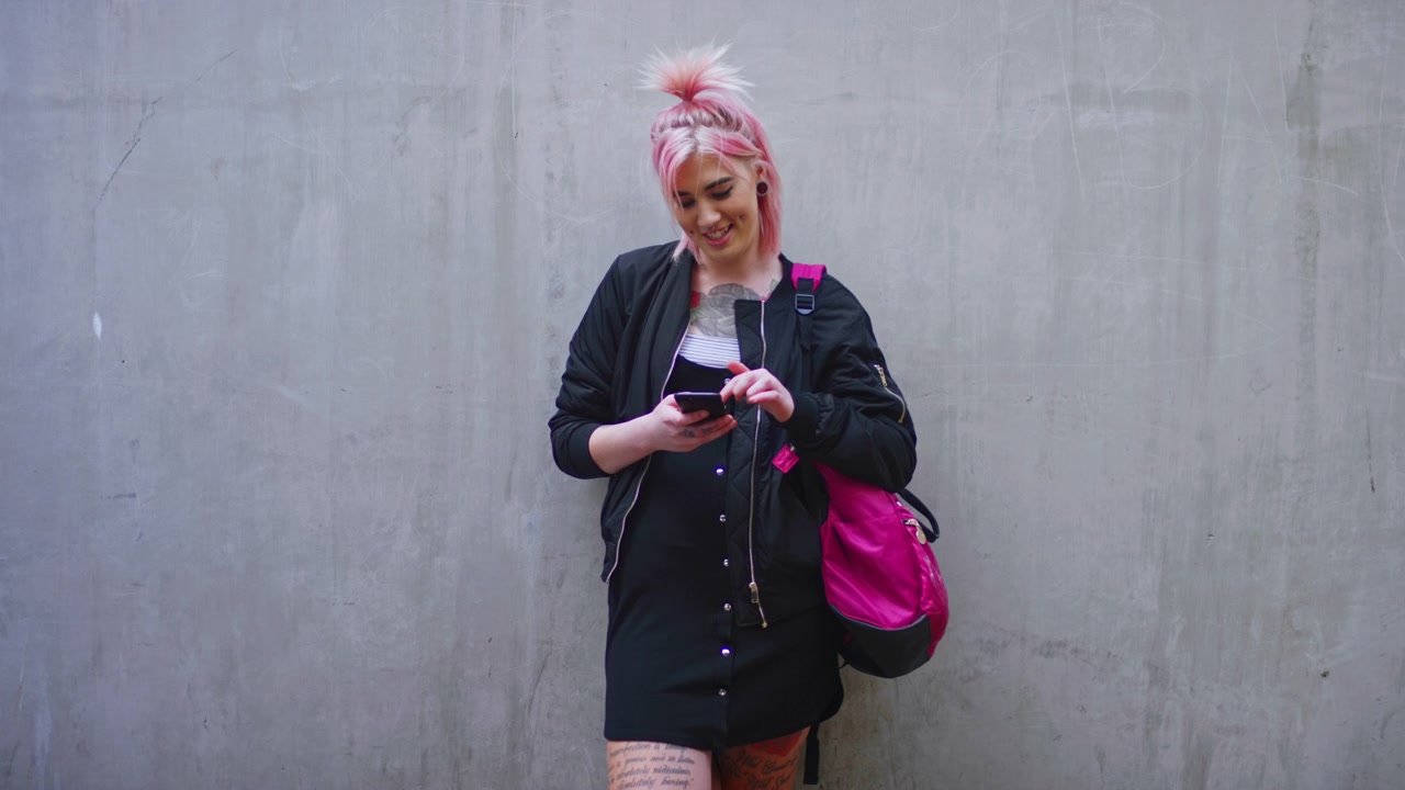 Giovane donna con capelli rosa che usa lo smartphone contro un muro di cemento