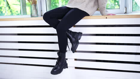 Model sitting on a white window sill showing the modern black boots with laces. Fashionable footwear demonstration on a female model.