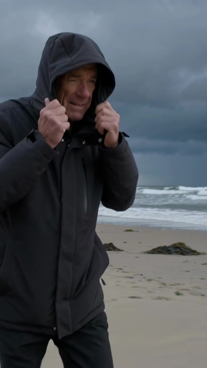 Vertical video: Pulling hood, senior walking on sandy shore with coat as storm coming, copy space