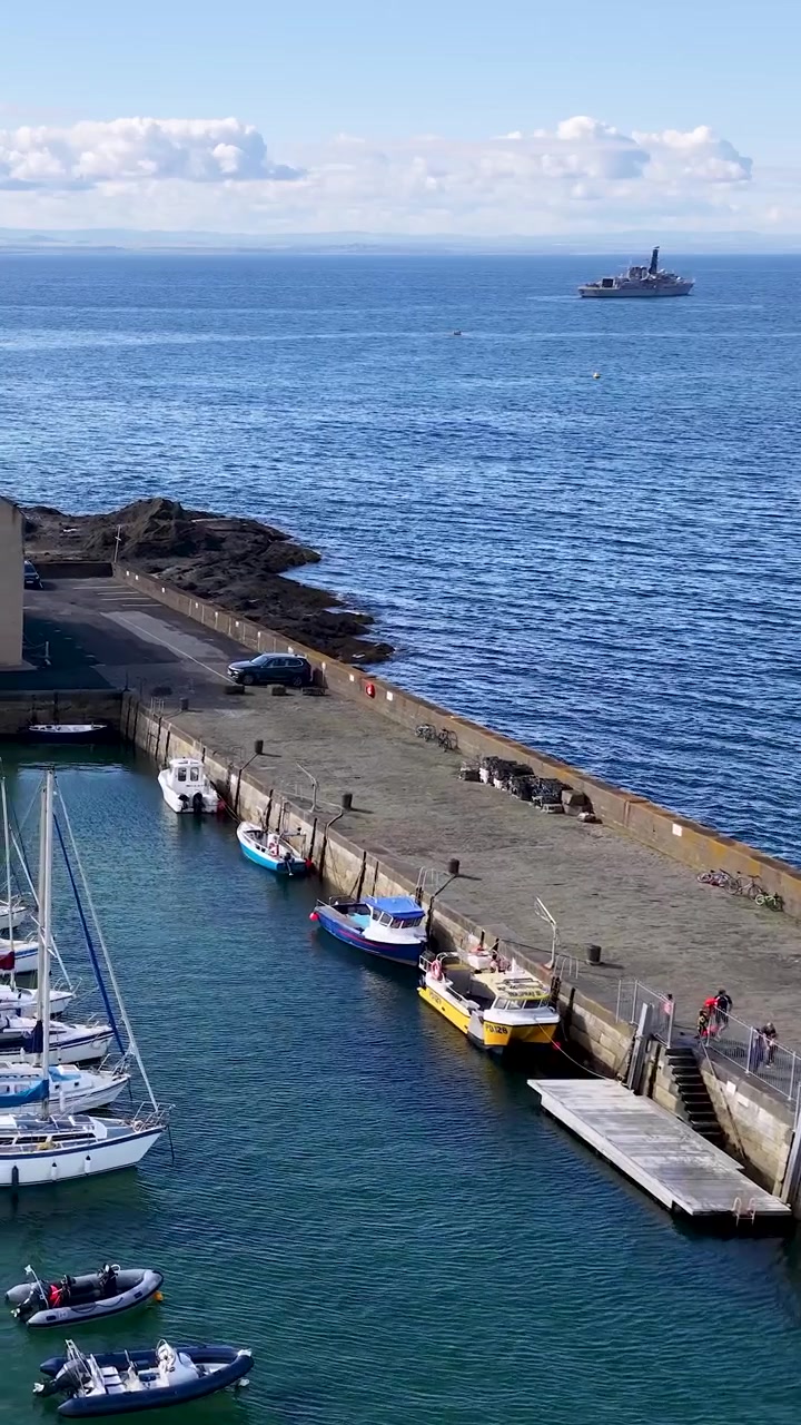 Un dron se desliza sobre el muelle de Fife, barcos y un puerto deportivo a plena luz del día con un mar azul en calma.