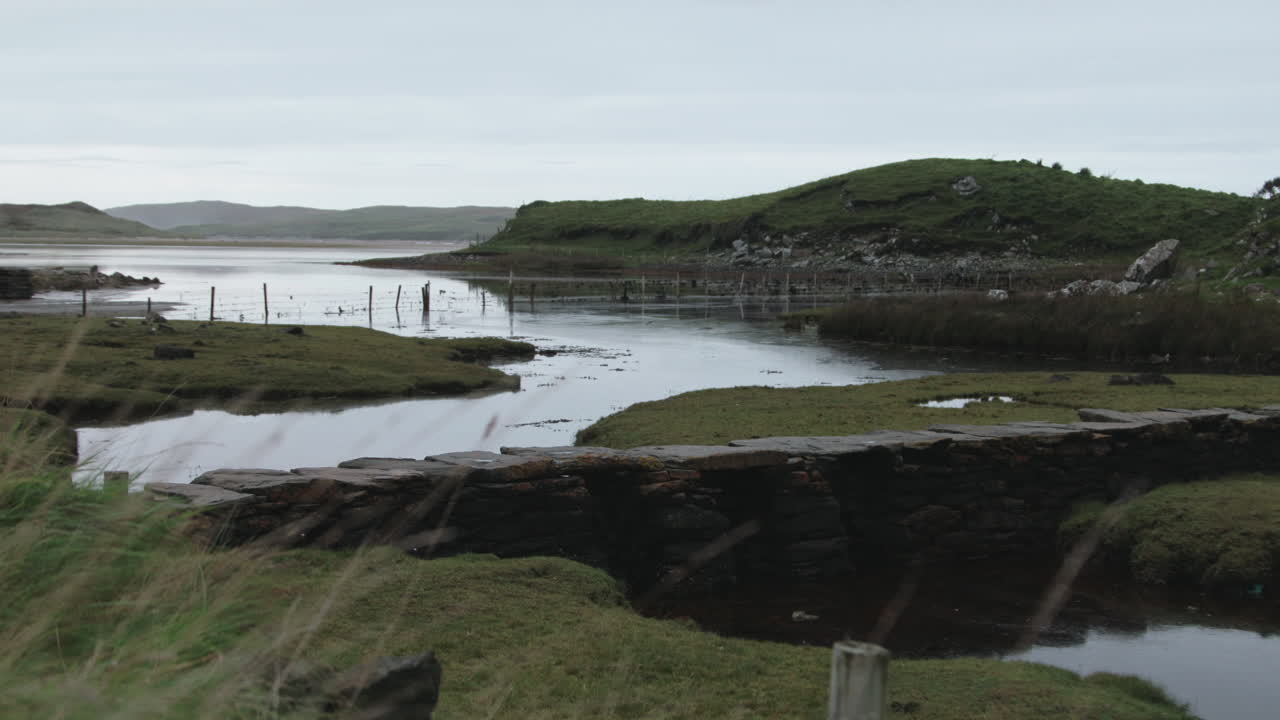 In North East Ireland's County Donegal, the early morning light casts a soft glow over still waters, surrounded by verdant grass and distant hills, creating a peaceful atmosphere.
