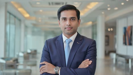 professional indian businessman wearing crisp blue suit, standing confidently with crossed arms in contemporary office lobby, representing corporate leadership and achievement