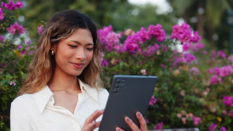 a woman happily using a tablet outdoors surrounded by vibrant flowers