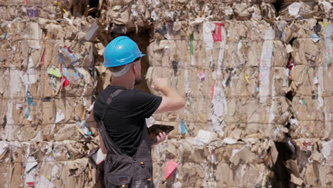 Recycling-plant-manager-with-blue-hard-hat-walks-and-counts-stacked-paper-bales