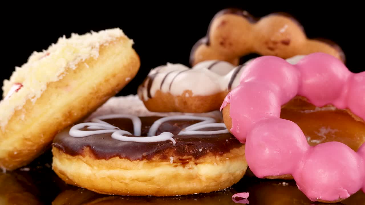 A variety of decorated donuts, including smiley face and chocolate glazed, rotate on a reflective surface with bright studio lighting and a black backdrop