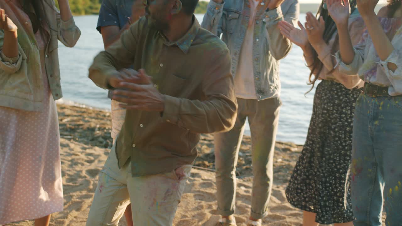 Joyful Middle Eastern man covered with gulal paint dancing with friends outdoors at beach party