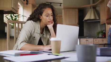 thoughtful biracial woman sitting in kitchen and working on laptop