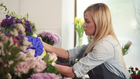 Young-attractive-blonde-female-florist-spraying-water-on-bunch-of-flowers-in-flower-shot.-Slow-Motion-shot