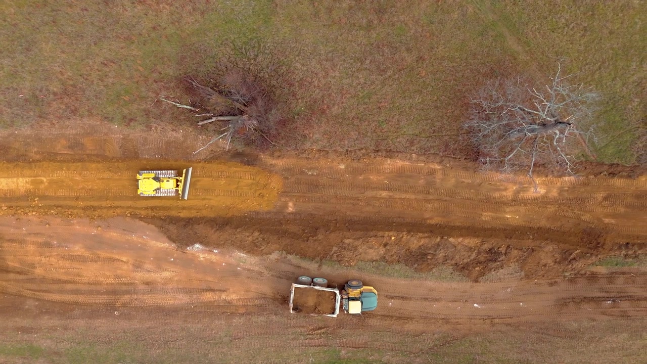 Aerial top down tracking follows a bulldozer leveling ground at a construction area as dump trucks drive parallel