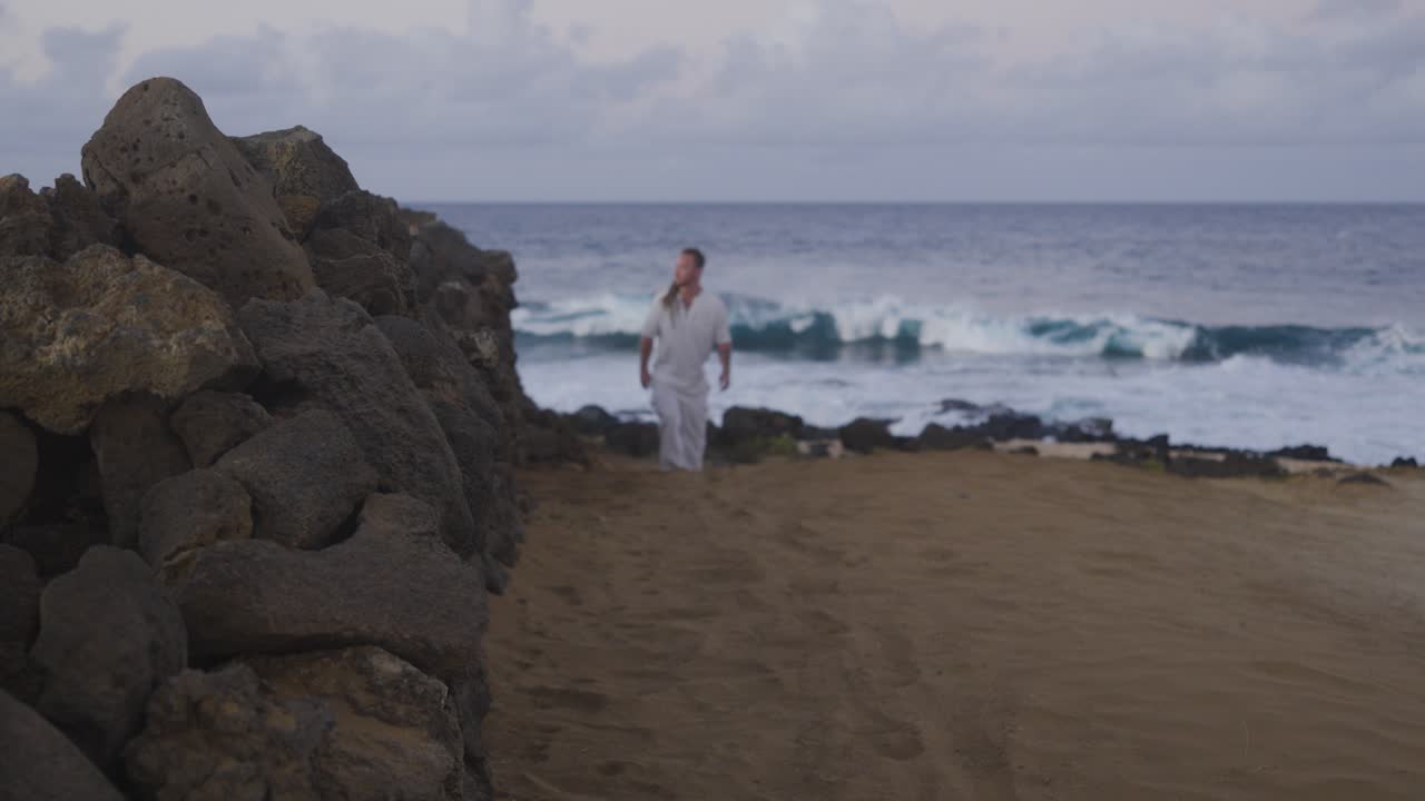 A wide view shows a man walking toward the camera on a sandy coastal path beside a rugged stone wall, with powerful ocean waves crashing in the background