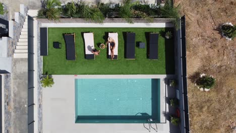 drone flies over an outdoor pool in bird's eye view - woman in swimming costume lies on sun lounger and man comes and sits next to her - villa in greece crete with palm trees