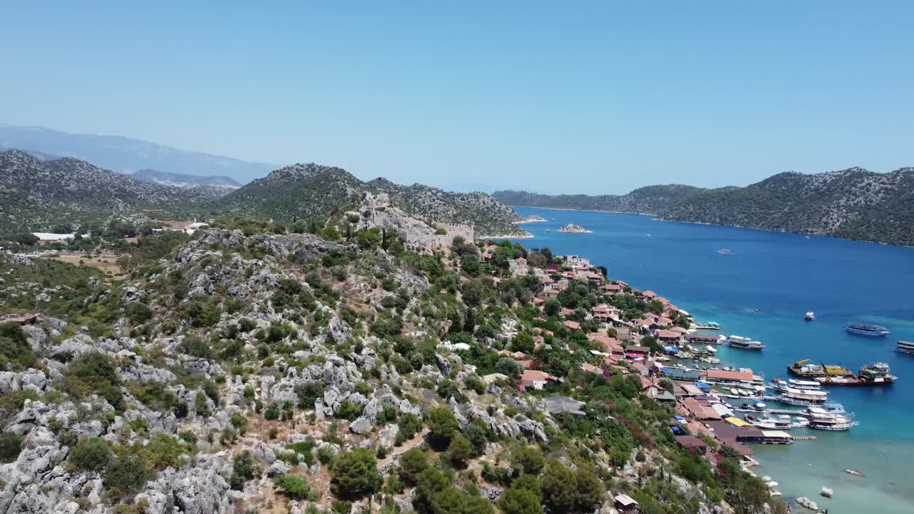 Aerial view of rugged hills beside a vibrant blue harbor. Scenic village structures dot the coast, creating a serene composition under clear skies in Kaş, Turkey. Captivating drone video