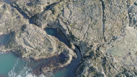 seguimiento aéreo sobre una costa rocosa, plana y desgastada, que revela tranquilas y vibrantes aguas marinas azules y pozas de rocas que brillan con reflejos del cielo.