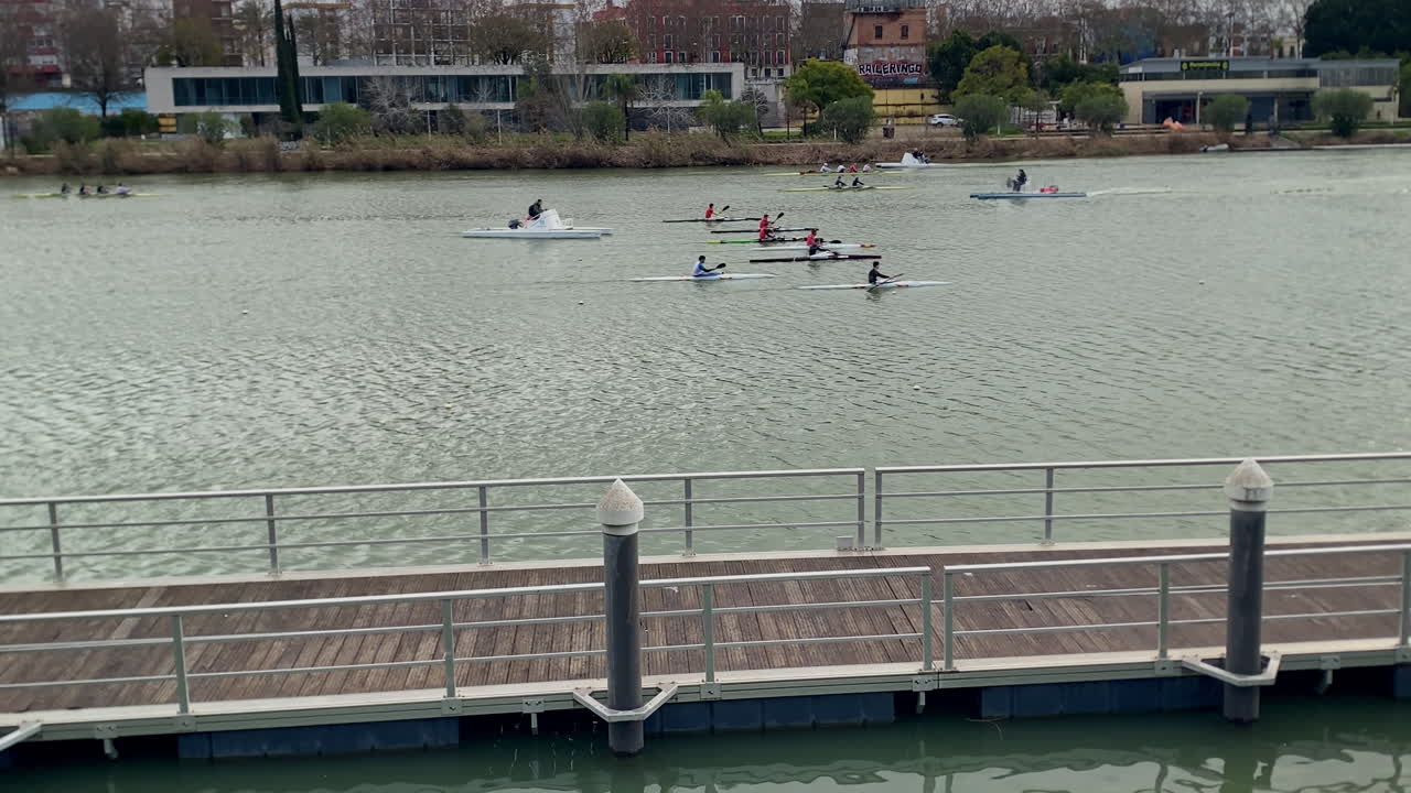 Atleter padler kajakker og kanoer under træning på Guadalquivir-floden i Sevilla, med byens bygninger på den modsatte bred.
