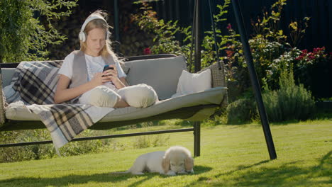 a teenage girl is resting on a garden swing, using a smartphone. her puppy sits next to her