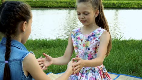 two happy little girls are sitting on the lawn by the river opposite each other and playing with their hands. they clap their hands laughing.