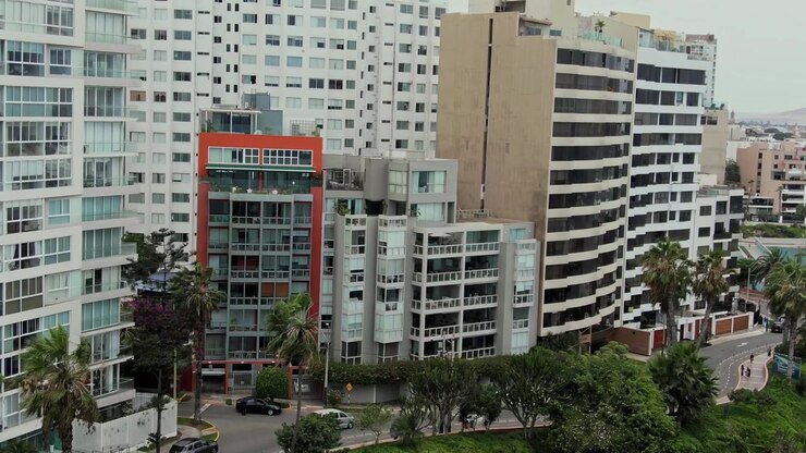 Vista aérea próxima das fachadas de apartamentos à beira-mar no Malecón de Miraflores em Lima, Peru, com varandas de vidro, terraços curvos, palmeiras e uma rua costeira tranquila