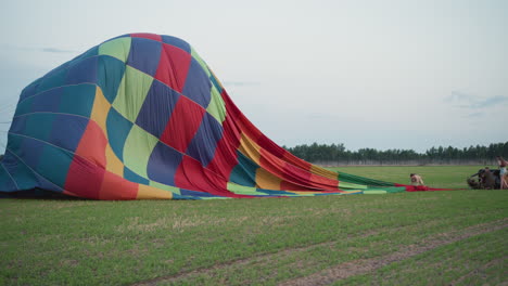 woman and man collapsing colorful hot air balloon envelope across green field at dusk while tethering rope and guiding canopy during controlled pack down operation under soft dusk sky in farmland