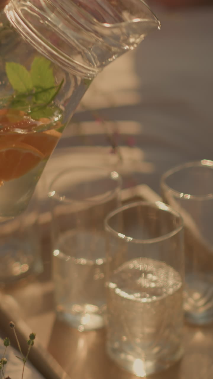 primer plano de la mano vertiendo agua refrescante infundida de la jarra con rebanadas de naranja y menta en vasos en bandeja de madera, bajo la cálida luz del sol, con vista parcial de la mesa de picnic