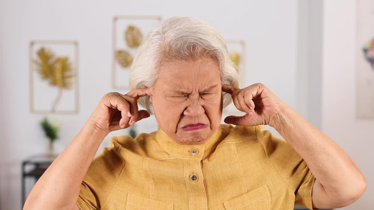 Senior Asian woman in yellow shirt covers her ears and winces, seated in a well-lit modern home interior with neutral decor and soft daylight