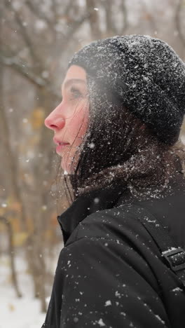 vista lateral de una dama con gorra negra y sudadera con capucha, salpicada de nieve, ajustando la correa de su paquete de guitarra mientras camina a través de un parque cubierto de nieve con árboles cargados de helada