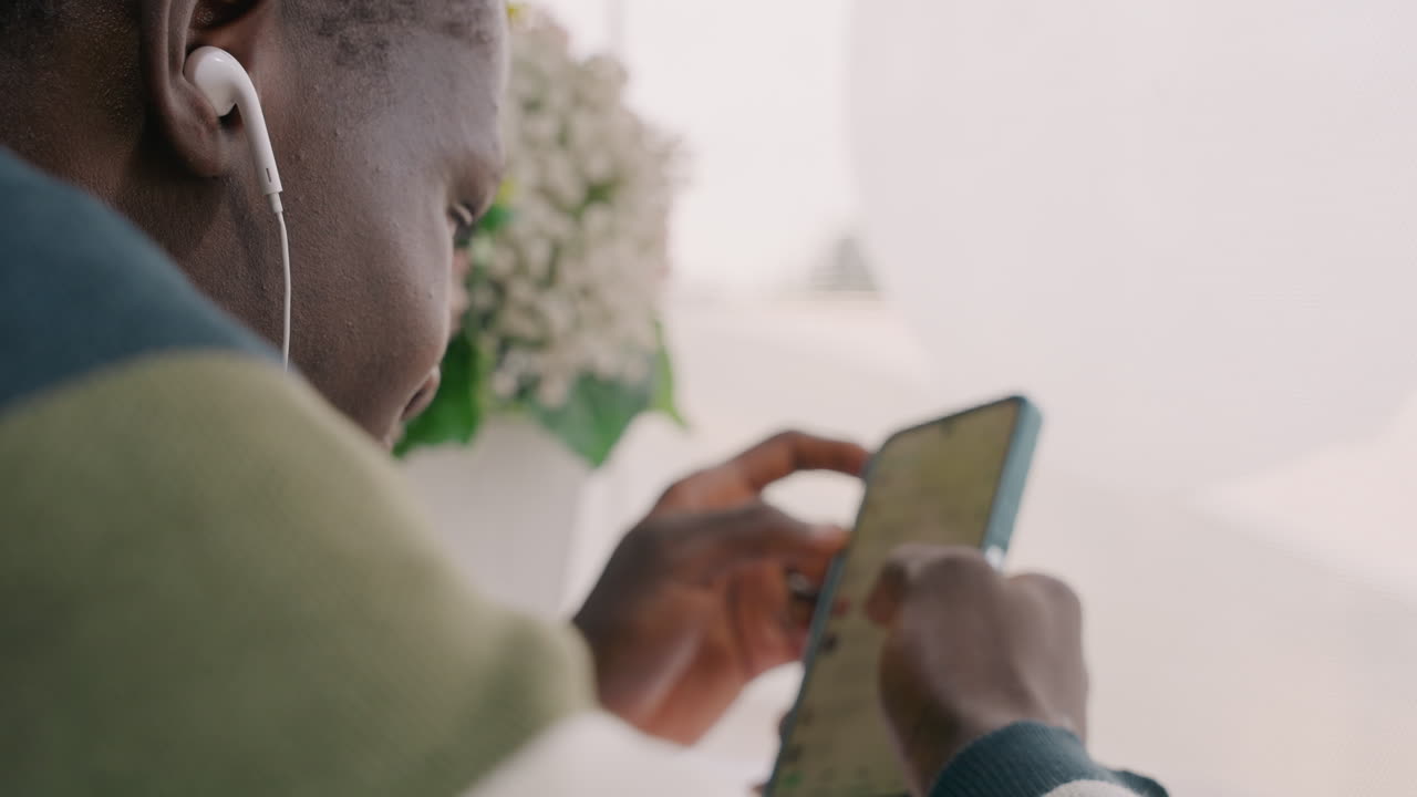 Black man texting on phone, earphones in ears, closeup of hands typing romantic message to partner during cozy cafe date, soft window light and floral decor, candid intimate mood with focused