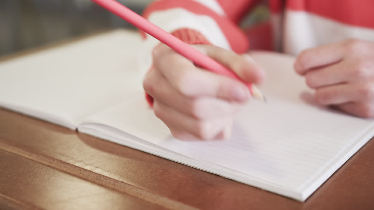 Young girl writing in notebook with pink pencil at school desk, focusing intently