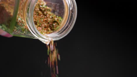 Static shot of falling delicious spices with basil, garlic, chili, paprika and pepper from a glass against black background