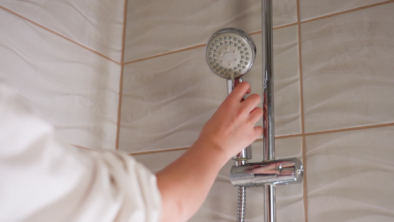 High angle view tiled bathroom shows chrome shower on wall holder as fair woman hand reaches, grips handle, keeps position for washing, light glints on rippled tiles, quiet indoor hygiene routine