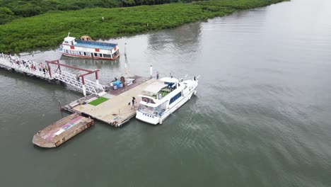 Volando-Un-Nuevo-Crucero-Fluvial-Pequeño-Atracado-En-Un-Embarcadero-En-El-Río-Con-Edificios-Y-Un-Puente-Rojo-En-Segundo-Plano-En-Un-Día-Nublado