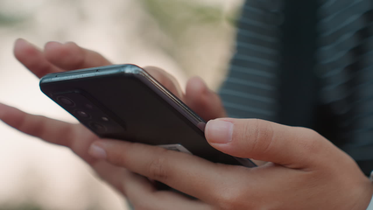 Primer plano de manos masculinas sosteniendo un smartphone al aire libre, con camisa a rayas y reloj visibles. Agarre firme mientras lee notificaciones y escribe con energía concentrada y profesional en un entorno urbano.