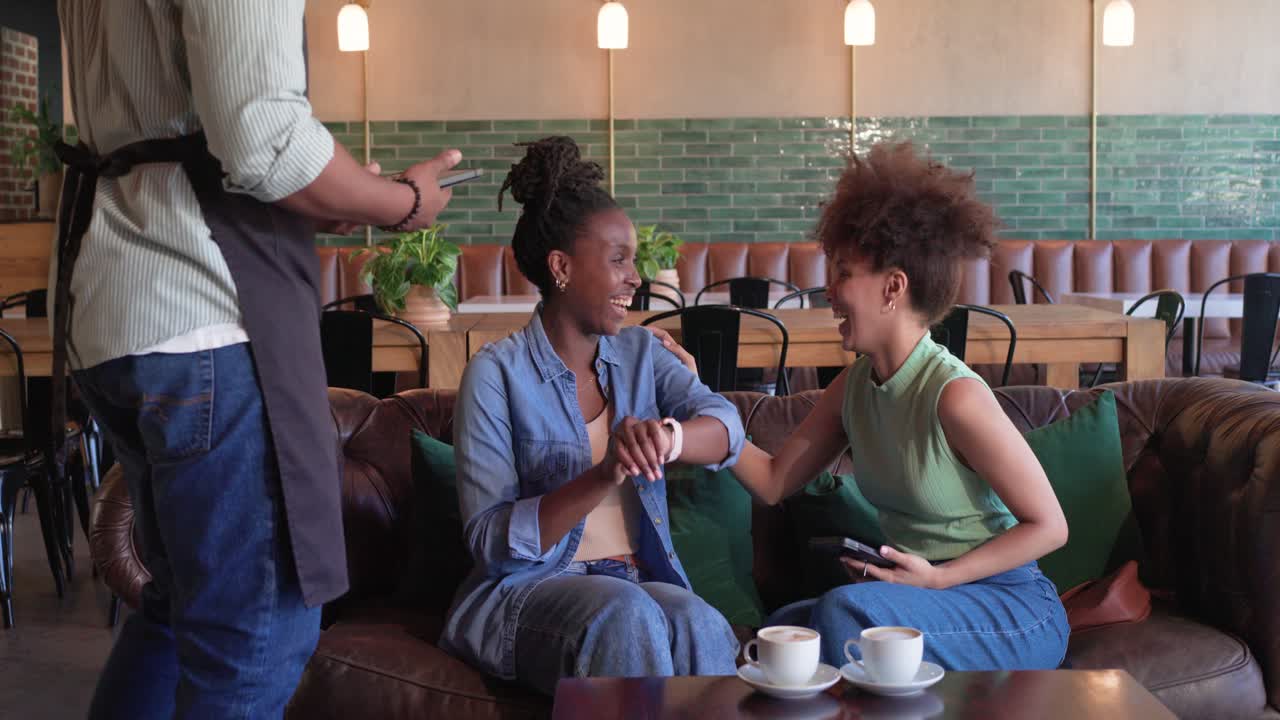 African American female friends comparing phones at cafe sofa while waiter showing tablet to order