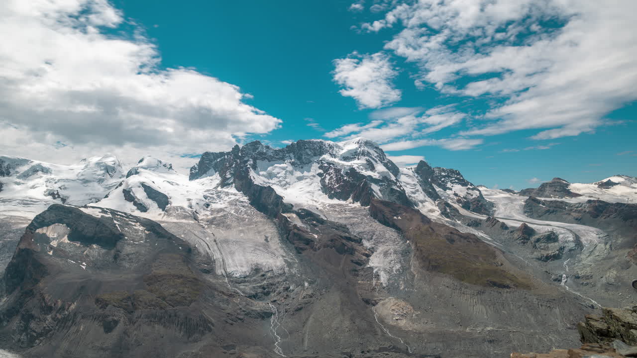 Timelapse of majestic snow-covered peaks under a bright blue sky. Glaciers and rocky ridges dominate the alpine scenery as clouds drift across the mountain range