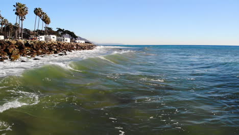 aerial drone shot flying low over ocean waves crashing on the rocky shore with palm trees and an rv campground on a california beach