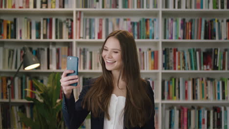 Retrato-De-Una-Joven-Hermosa-Mujer-Parada-En-La-Biblioteca-Usando-Un-Teléfono-Inteligente-Video-Chat-Estudiante-Saludando-Sonriendo-Riendo