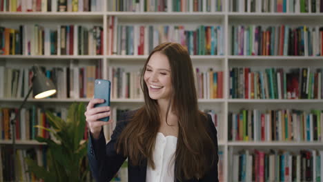 Porträt-Einer-Jungen-Schönen-Frau,-Die-In-Der-Bibliothek-Steht-Und-Einen-Smartphone-Video-Chat-Nutzt.-Studentin-Winkt,-Lächelt-Und-Lacht