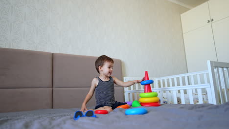 male kid sits on the bed playing with a pyramid. baby boy plays with a toy in the bedroom.