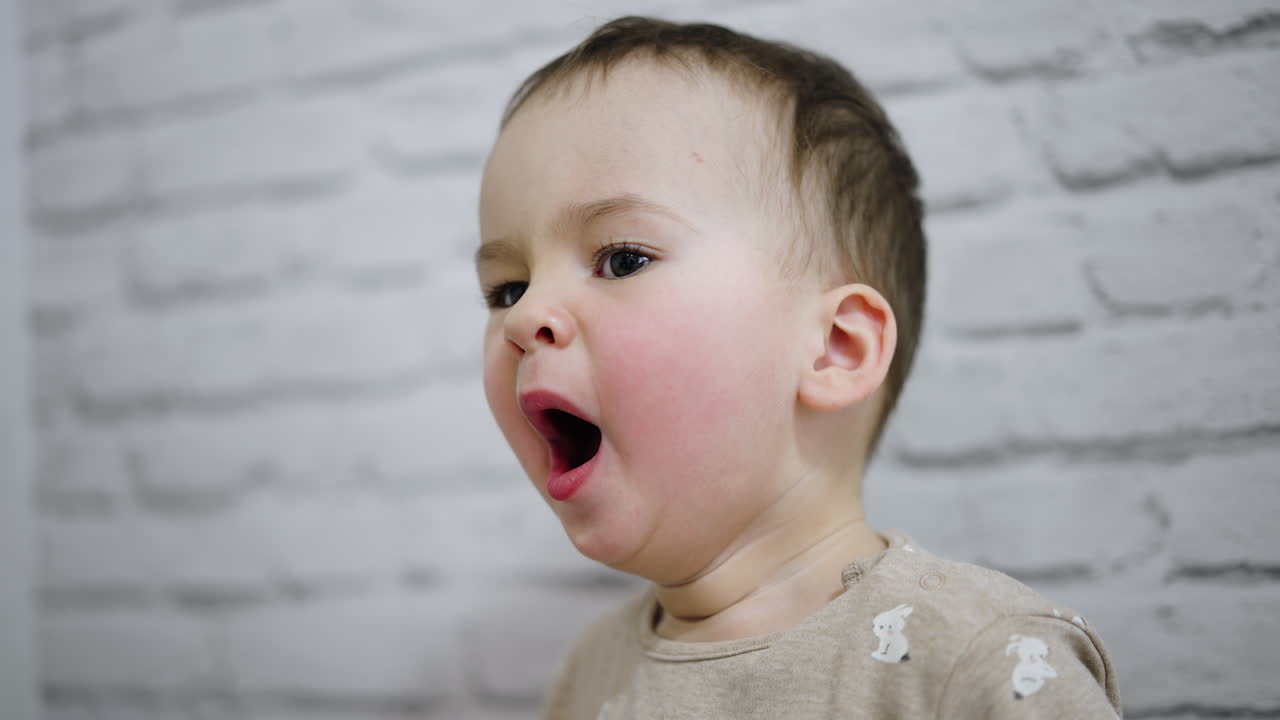 Charming baby boy with rosy cheeks yawning in front of camera. White backdrop.
