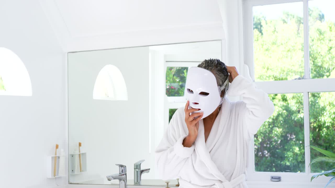 Senior African woman in white robe fitting mask pulling strap at vanity mirror preparing skincare