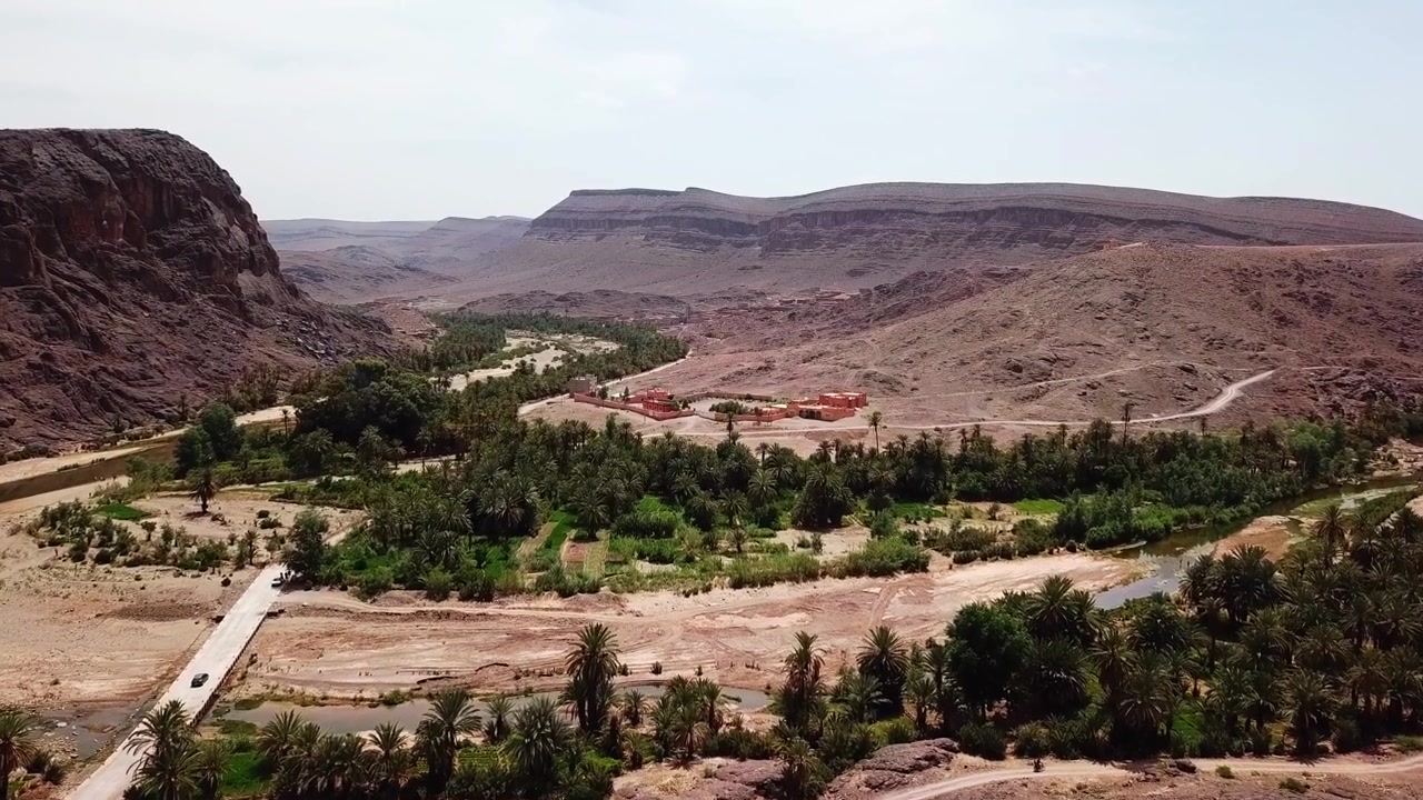 Filmagens aéreas cinematográficas de drone de um vale oásis com estradas sinuosas e vegetação verde, rodeado por montanhas desérticas em Ouarzazate, Marrocos. Ideal para produções de viagem, documentário e cinema.